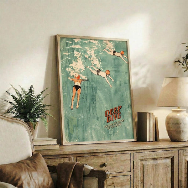 May include: Vintage-style poster with turquoise background showing three divers in mid-dive poses, red text reading "DEEP DIVE" at bottom. Framed artwork displayed on rustic wood sideboard next to fern plant, ceramic vase, and books in cozy living room setting with beige armchair and neutral walls.