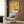 May include: Mid-century modern living room with beige upholstered armchair featuring geometric patterned throw pillow. Yellow abstract art poster with black organic shapes, crescent moon, and orange accent mounted on wood-paneled wall. Gold brass floor lamp with white circular base stands beside chair. Dark round side table holds open book. Warm neutral tones throughout with natural lighting from window.