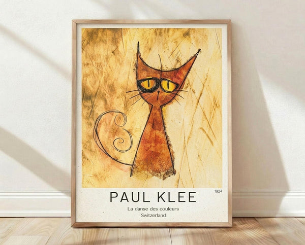 May include: Framed Paul Klee art print featuring stylized orange cat with pointed ears, yellow eyes, and curly tail on textured golden background. Poster displays "PAUL KLEE 1924 La danse des couleurs Switzerland" text below artwork. Light wood frame with white matting, leaning against white wall on wooden floor with soft natural lighting and shadows.