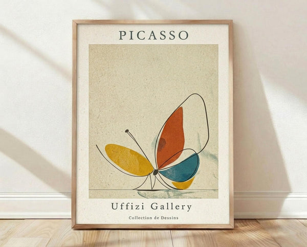 May include: Framed poster print featuring an abstract butterfly illustration in Picasso's minimalist style, with colorful geometric wings in yellow, orange, and teal against a cream textured background. The artwork shows "PICASSO" at top and "Uffizi Gallery Collection de Dessins" at bottom in dark text. Natural wood frame sits on light wooden floor against white textured wall with soft natural lighting casting shadows.