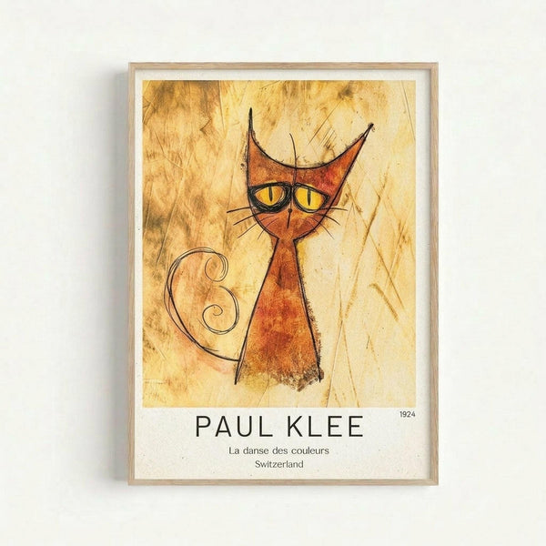 Framed art print featuring a stylized orange cat with pointed ears, yellow eyes, and curly tail on aged parchment background. The modernist illustration uses warm earth tones with black line work. Below the artwork is text reading "PAUL KLEE" with "La danse des couleurs Switzerland 1924". The print is displayed in a light wooden frame with white matting against a neutral wall.