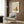 May include: Framed poster with Picasso and Uffizi Gallery text featuring abstract colorful butterfly design in orange, yellow, and teal on cream background, displayed on wood paneled wall above beige upholstered chair with geometric patterned pillow, brass floor lamp, and dark round side table with open book in modern living room setting