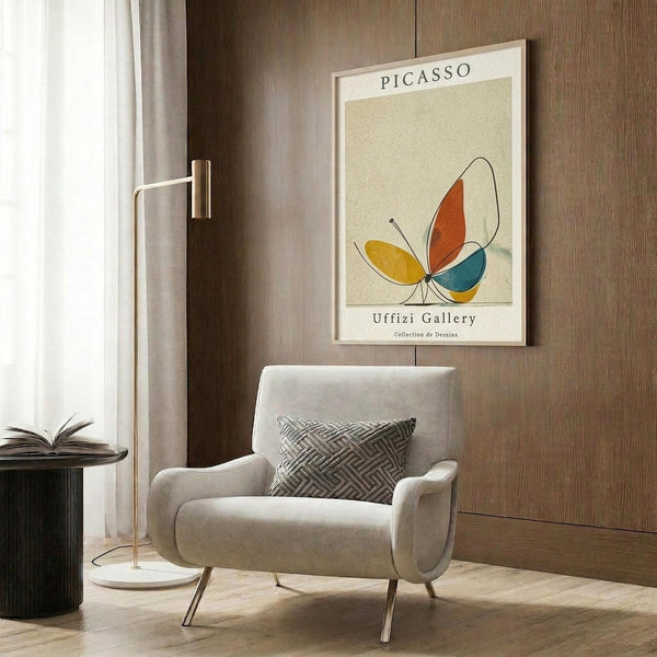 May include: Framed poster with Picasso and Uffizi Gallery text featuring abstract colorful butterfly design in orange, yellow, and teal on cream background, displayed on wood paneled wall above beige upholstered chair with geometric patterned pillow, brass floor lamp, and dark round side table with open book in modern living room setting