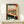 May include: Framed Japanese woodblock print showing interior view through wooden window bars overlooking landscape with Mount Fuji. White cat sits on windowsill beside folded cloth. Traditional tatami room with geometric floor patterns in green and black. Artwork features classic ukiyo-e style with red cartouches containing Japanese text. Small wrapped gift box sits on light wooden shelf below the gold-framed print against white wall.