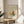 May include: Framed poster featuring a minimalist butterfly design with colorful wings in yellow, orange, and teal on beige background, displayed on a wooden round side table next to a beige upholstered chair. Sunlight streams through white curtains creating shadows on the neutral wall. A green plant leaf is partially visible in the corner.