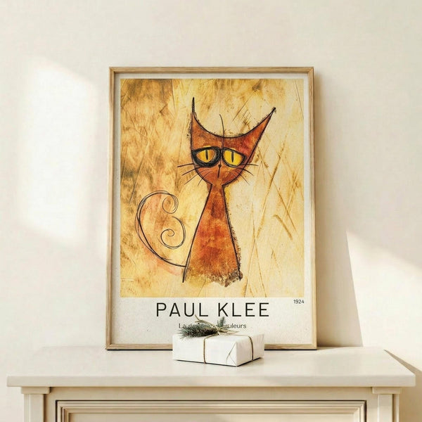 May include: Framed art print featuring an abstract orange cat with pointed ears, yellow eyes, and curved tail on aged paper background, displayed on light wood shelf next to small white gift box with greenery sprig against white wall.