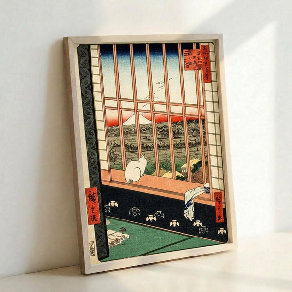Framed reproduction of a traditional Japanese woodblock print showing a white cat sitting on a windowsill, looking out through wooden lattice bars at Mount Fuji in the distance. The interior features tatami mats with floral patterns in green and black, while the outdoor view shows a pastoral landscape with fields and mountains under a gradient sky. Red Japanese text cartouches appear in the corners. The print is displayed in a simple wooden frame against a white wall.