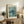May include: Framed art print featuring a detailed sea turtle swimming among coral-like vegetation in muted blue-green tones, displayed on a rustic wooden sideboard alongside books, a ceramic vase, and fern plant in a cozy living room setting with cream walls and brown leather furniture