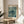 May include: Framed poster featuring a sea turtle swimming among coral and seaweed in muted green and teal tones, displayed on a round wooden side table next to a beige upholstered chair. Natural sunlight streams across the white walls, with a plant's leaves visible in the foreground. The artwork shows text reading "YAYOI KUSAMA" at top and "Modern Art 1966" at bottom in a light wooden frame.