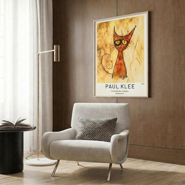 May include: Framed poster featuring Paul Klee's stylized orange and red cat with yellow eyes on aged parchment background, displayed on wood paneled wall above beige upholstered armchair with geometric patterned pillow, brass floor lamp, and dark round side table with open book in modern neutral living room setting.