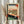 May include: Hands holding a framed Japanese woodblock print showing a white cat sitting by a window overlooking a landscape with Mount Fuji. The artwork features traditional ukiyo-e style with wooden window frames, tatami mats, and red Japanese text cartouches. Dried pampas grass visible to the left. The print is displayed in a light wooden frame against white curtains in soft natural lighting.