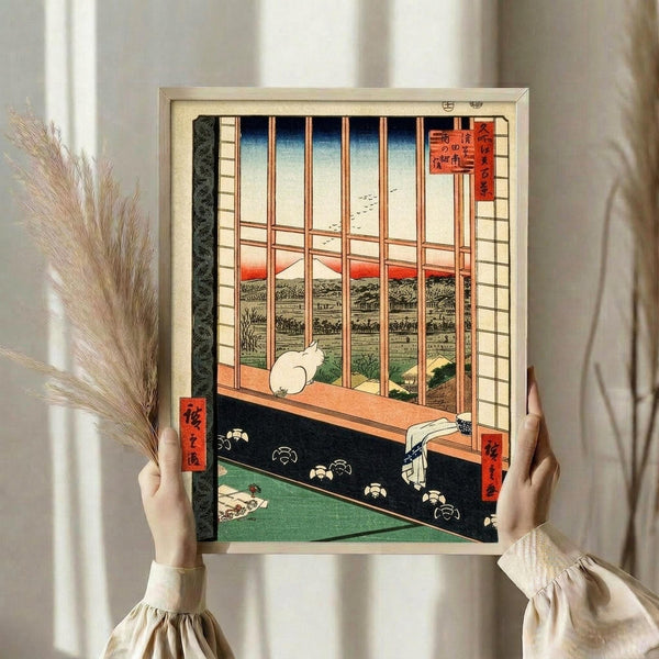 May include: Hands holding a framed Japanese woodblock print showing a white cat sitting by a window overlooking a landscape with Mount Fuji. The artwork features traditional ukiyo-e style with wooden window frames, tatami mats, and red Japanese text cartouches. Dried pampas grass visible to the left. The print is displayed in a light wooden frame against white curtains in soft natural lighting.