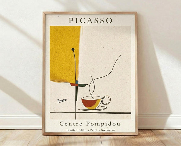 May include: Framed abstract art print in light wood frame leaning against white wall on wooden floor. Artwork features minimalist line drawing of coffee cup with steam on cream background, yellow geometric rectangle, red and green accents. Print shows "PICASSO" at top, "Centre Pompidou" and "Limited Edition Print No. 04/50" at bottom with signature. Soft natural lighting creates diagonal shadows across white textured wall.