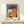 May include: Framed Bauhaus poster with geometric orange, blue, and cream rectangular shapes on dark blue background, displayed on light wood console table next to small wrapped gift with greenery accent against cream wall