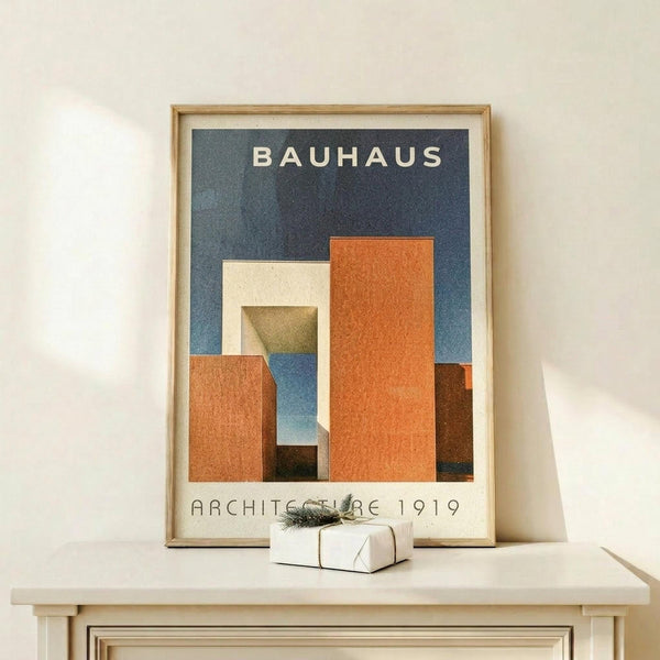 May include: Framed Bauhaus poster with geometric orange, blue, and cream rectangular shapes on dark blue background, displayed on light wood console table next to small wrapped gift with greenery accent against cream wall