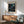 May include: Framed poster with cosmic swirling patterns in turquoise and navy blue against black background, featuring scattered dots and celestial orbs. Displayed above mid-century modern wooden credenza with white ceramic lamp. Sage green upholstered armchair with wooden frame sits nearby on wooden floor. Soft natural light creates shadows on white wall.