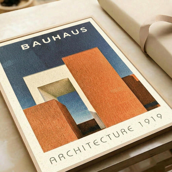 Vintage-style poster featuring geometric architectural forms in orange, blue, and cream colors with "BAUHAUS" text at top and "ARCHITECTURE 1919" at bottom. The design shows overlapping rectangular shapes creating an abstract building composition. Print is displayed on cream paper with rolled paper tubes visible in the background, showing fine texture and matte finish.