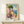 May include: Framed vintage-style travel poster showing colorful Mediterranean coastal village with pink and yellow buildings, large green tree, blue water, and Italian text at bottom. Displayed on light wood shelf with small wrapped gift box beside it against white wall.