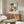 May include: Framed vintage-style poster featuring colorful Mediterranean coastal scene with pastel buildings, leafy tree, and blue water, displayed above wooden sideboard in modern living room with white upholstered chair, glass coffee table, and neutral decor