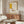 May include: Mid-century modern living room with cream walls featuring a gold-framed art print displaying abstract yellow rectangle, teacup, and geometric lines in Picasso's style with "Centre Pompidou" text. Scene shows walnut wood sideboard, white curved lounge chair, glass-top coffee table, and minimalist table lamp creating warm, sophisticated interior styling.