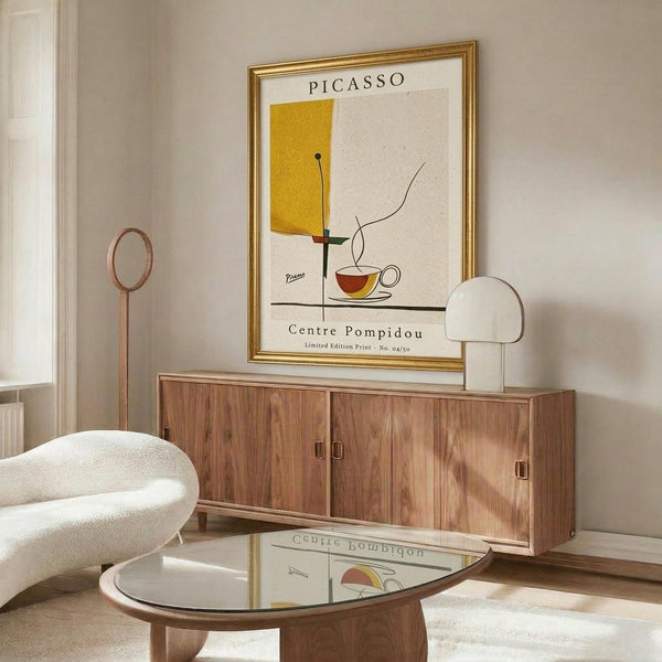 May include: Mid-century modern living room with cream walls featuring a gold-framed art print displaying abstract yellow rectangle, teacup, and geometric lines in Picasso's style with "Centre Pompidou" text. Scene shows walnut wood sideboard, white curved lounge chair, glass-top coffee table, and minimalist table lamp creating warm, sophisticated interior styling.