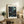 May include: Framed poster with cosmic swirling patterns in deep blues and teals with scattered white and yellow dots, displayed on rustic wood sideboard. Room scene shows neutral armchair, potted fern, ceramic vessels, and books creating cozy living space with cream walls and warm natural lighting.