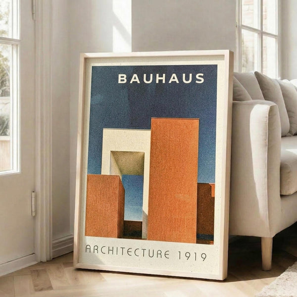 May include: Framed poster with blue background featuring geometric orange, cream, and brown architectural shapes. White text reads "BAUHAUS" at top and "ARCHITECTURE 1919" at bottom. Light wood frame leans against white wall next to beige sofa in bright, minimalist living room with large windows and natural lighting.