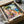 May include: Colorful Italian coastal village poster print featuring Mediterranean houses in pink, orange and yellow tones with a large tree, blue water with sailboat in background. Print shown in black frame inside cardboard shipping box with protective tissue paper wrapping.