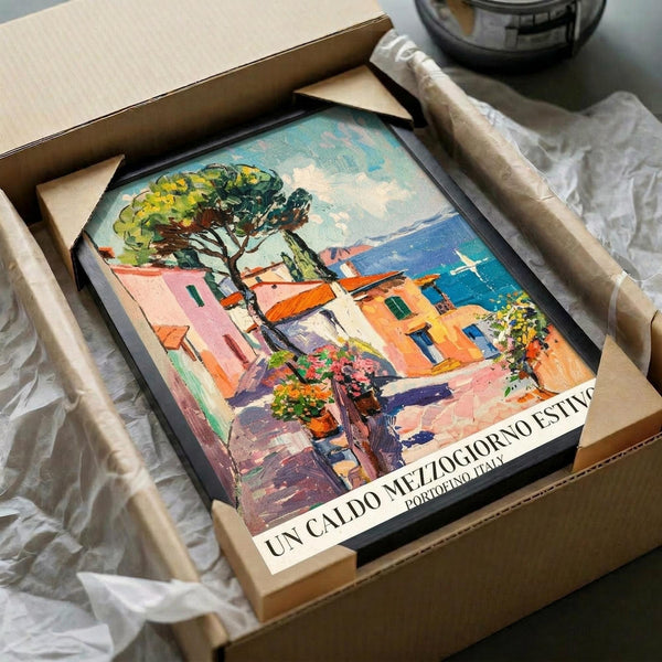 May include: Colorful Italian coastal village poster print featuring Mediterranean houses in pink, orange and yellow tones with a large tree, blue water with sailboat in background. Print shown in black frame inside cardboard shipping box with protective tissue paper wrapping.