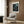 May include: Framed art poster with cosmic design featuring swirling blue and green patterns, white dots and circles on dark background, displayed above modern cream armchair with geometric pillow. Room has wood panel walls, brass floor lamp, and dark round side table with open book.