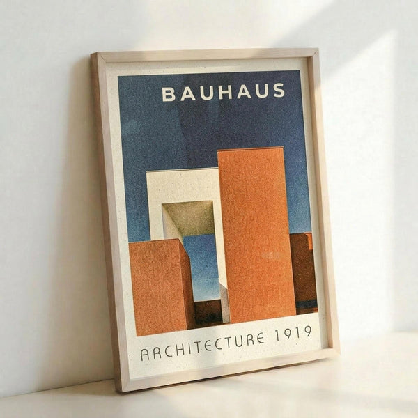 Framed poster with geometric architectural forms in orange, cream, olive, and brown against a dark blue background. White text reads "BAUHAUS" at top and "ARCHITECTURE 1919" at bottom. Light wood frame with white matting, displayed against white wall corner.