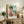 May include: Framed vintage-style travel poster showing colorful Mediterranean coastal village with pink and yellow buildings, large green tree, and blue ocean. Displayed on rustic wooden sideboard with books, ceramic vase, and fern plant in neutral-toned living room setting.