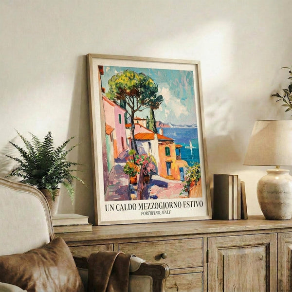 May include: Framed vintage-style travel poster showing colorful Mediterranean coastal village with pink and yellow buildings, large green tree, and blue ocean. Displayed on rustic wooden sideboard with books, ceramic vase, and fern plant in neutral-toned living room setting.