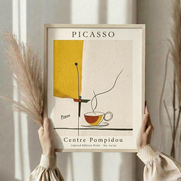 May include: Person holding framed minimalist abstract art print in light wood frame. Artwork features yellow geometric shape, red and yellow coffee cup, and thin black lines on cream background with "PICASSO" and "Centre Pompidou" text. Styled with dried pampas grass against soft neutral curtained background.