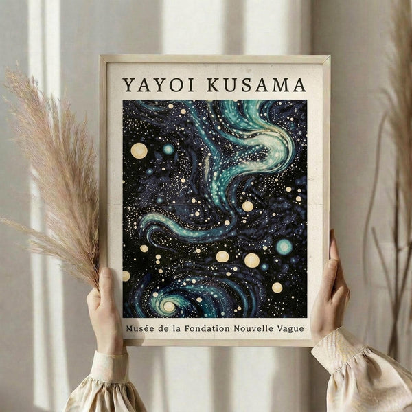 May include: Hands holding a framed art print featuring swirling cosmic patterns in deep blue and teal with scattered dots and celestial orbs, alongside dried pampas grass against a cream curtained background. The artwork shows flowing, galaxy-like formations with white and yellow circular elements throughout the composition.