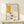 May include: Framed Picasso-style abstract art print featuring yellow geometric shape, teacup with steam, and minimalist line drawing on cream background. Natural wood frame displayed on light wooden shelf beside small wrapped gift with pine sprig against white wall.