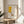 May include: Framed abstract art print in light wood frame displayed on round wooden side table next to beige upholstered chair. Print features yellow geometric shape, stylized coffee cup in red and yellow, and black linear elements on cream background with "PICASSO" and "Centre Pompidou" text. Soft natural lighting creates shadows on white wall, with plant leaves visible in corner.