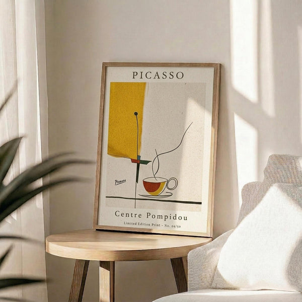 May include: Framed abstract art print in light wood frame displayed on round wooden side table next to beige upholstered chair. Print features yellow geometric shape, stylized coffee cup in red and yellow, and black linear elements on cream background with "PICASSO" and "Centre Pompidou" text. Soft natural lighting creates shadows on white wall, with plant leaves visible in corner.
