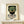 May include: Framed vintage-style poster featuring an Aztec mosaic mask in green turquoise tiles with red geometric patterns and white eyes, displayed on a light wooden shelf next to a small wrapped gift, against a cream wall in modern interior setting