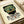 May include: Framed art print featuring a stylized green mosaic-patterned mask with red geometric accents and white eyes, displayed in a light wooden frame on cream paper. Spanish text below reads "EL MAESTRO" with publication details including Mexico and Roman numerals. The print has a vintage poster aesthetic with earthy tones, shown alongside rolled paper tubes on a light wooden surface.