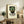May include: Framed vintage-style poster featuring mosaic mask face in green tiles with red accents and Spanish text, displayed on rustic wooden sideboard with books, ceramic vase, and fern plant in neutral-toned living room setting