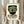 May include: Framed vintage-style poster with cream background featuring green mosaic mask design with red geometric patterns and white eyes. Person in beige ruffled sleeves holds wooden frame next to dried pampas grass against soft curtained window backdrop.