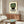 May include: Framed vintage-style poster featuring green mosaic mask with geometric patterns and red accents on cream background with Spanish text, displayed above walnut credenza in modern living room with white curved chair and oval glass coffee table