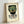 May include: Framed vintage-style poster leaning against white wall featuring an Aztec mosaic mask in green and red tiles with geometric patterns. The mask has white eyes and is set against black background. Spanish text below reads "EL MAESTRO REVISTA DE CULTURA NACIO NAL ENERO FEBRERO MEXICO MCMXXII" in bold black letters. Light wood frame with cream textured matting creates museum-quality presentation.