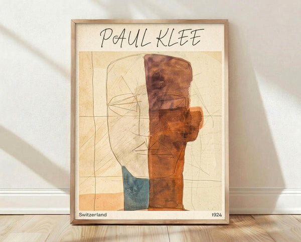 May include: Framed Paul Klee abstract artwork print displayed against white wall on wooden floor. The poster features geometric shapes in warm oranges, browns, and blue tones with linear pencil drawings on aged cream paper background. Light wooden frame surrounds the vintage-style reproduction with "Switzerland 1924" text at bottom. Natural lighting creates soft shadows on the wall.