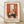 May include: Framed vintage-style poster print leaning against white wall on light wooden floor. Features orange and cream artwork showing three cats around a black round table with green vase containing flowering branches, set near a window. French text at bottom. Natural lighting creates soft shadows on textured wall.