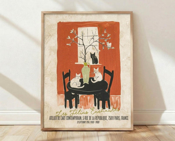 May include: Framed vintage-style poster print leaning against white wall on light wooden floor. Features orange and cream artwork showing three cats around a black round table with green vase containing flowering branches, set near a window. French text at bottom. Natural lighting creates soft shadows on textured wall.