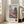 May include: Framed poster print leaning against white wall next to beige sofa in bright living room. Artwork shows Japanese pagoda temple with pink cherry blossom trees in vintage illustration style. Features Japanese text on left side, "SAKURA" text at bottom, and red circular symbol. Light wood frame, cream matting, positioned on wooden floor near large windows.