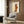 May include: Modern living room with beige armchair featuring geometric patterned pillow, brass floor lamp, dark round side table with open book, and framed abstract art poster on wood-paneled wall. The artwork shows warm orange and rust colored geometric shapes with linear elements on cream background in light wooden frame.