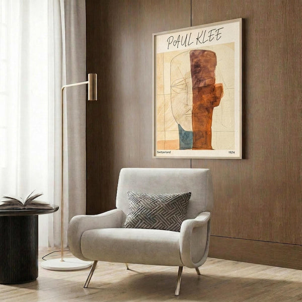 May include: Modern living room with beige armchair featuring geometric patterned pillow, brass floor lamp, dark round side table with open book, and framed abstract art poster on wood-paneled wall. The artwork shows warm orange and rust colored geometric shapes with linear elements on cream background in light wooden frame.